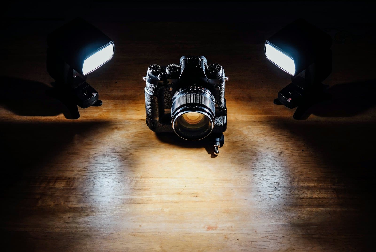 A camera between two lit studio flashes on a wooden table, seen from above